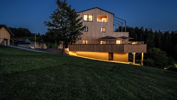 A modern two-story house with a sloped roof, large windows, and a well-lit exterior.