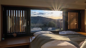 Een kamer met een bed, een raam met zonwering en uitzicht op een bebost landschap.