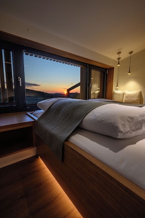 A bedroom with a large window, a bed, bedside tables, and a nightstand. - Modern design apartment with a view of the Dreisesselberg.<br>With sauna, roof terrace, outdoor bathtub and custom-made furnishings.<br>Perfect for two adults who appreciate peace and quality. (Breitenberg)