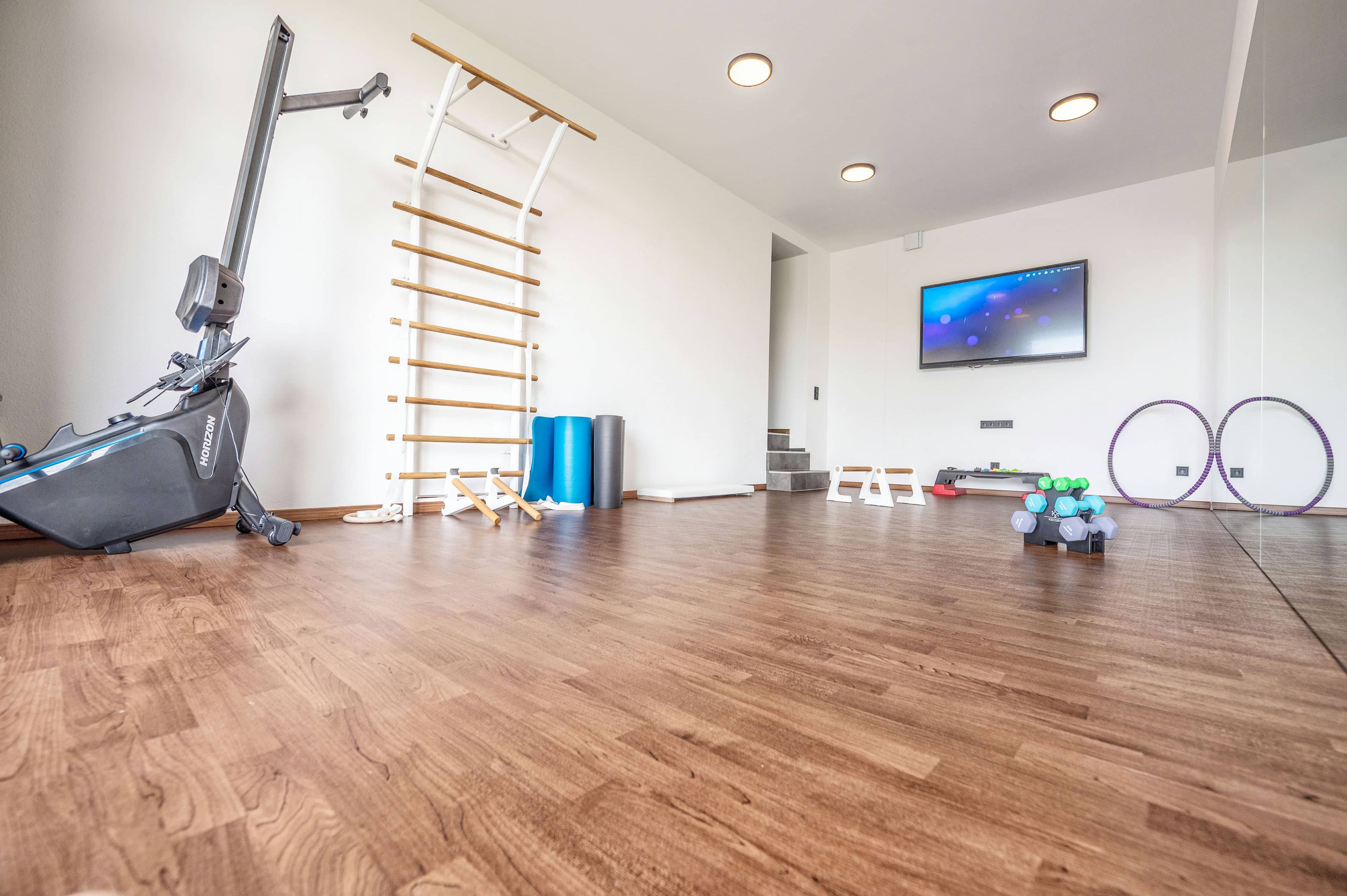 A spacious room with a wooden floor, a ceiling-mounted exercise machine, a wall-mounted TV, and a yoga mat.