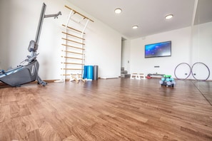 A spacious room with a wooden floor, a ceiling-mounted exercise machine, a wall-mounted TV, and a yoga mat.