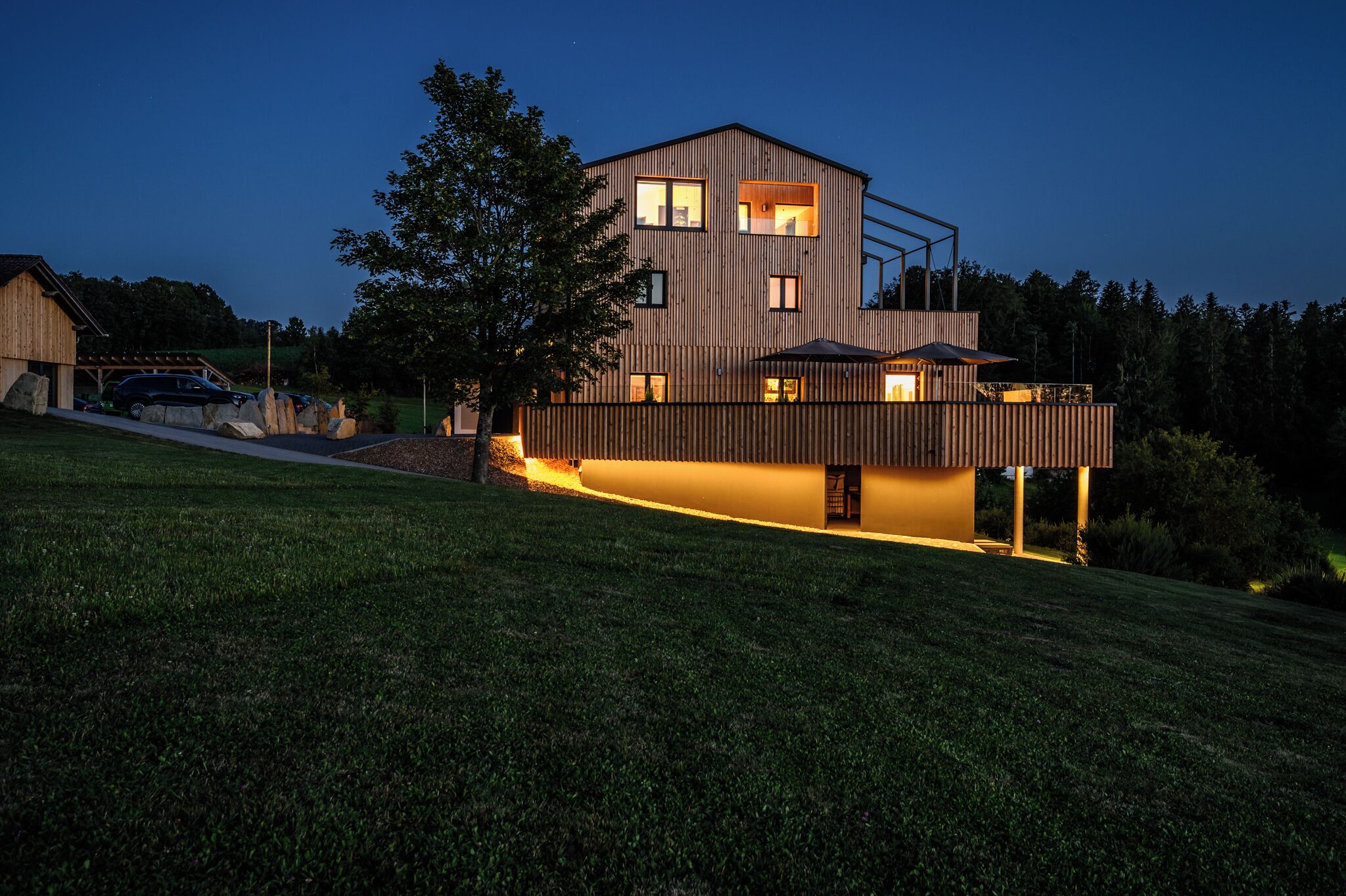A modern two-story house with a sloped roof, large windows, and a well-lit exterior.