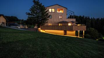 A modern two-story house with a sloped roof, large windows, and a well-lit exterior.