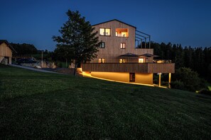 Une maison moderne à deux étages, dotée d’un toit en pente, de grandes fenêtres et d’un éclairage extérieur soigné.