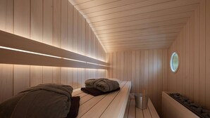 A sauna room with wooden walls and benches, a small circular window, and a trash bin. - Modern design apartment with a view of the valley.<br>With sauna, roof terrace, outdoor bathtub and custom-made furnishings.<br>Perfect for two adults who appreciate peace and quality. (Breitenberg)
