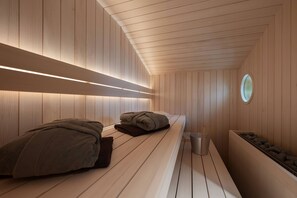 A sauna room with wooden walls and benches, a small circular window, and a trash bin.