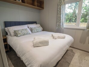 A neatly made bed with white bedding and floral pillows, a wooden headboard, a window with curtains, and a small wooden shelf above the bed. - The Oaks 2 Bedrooms (Northwich)