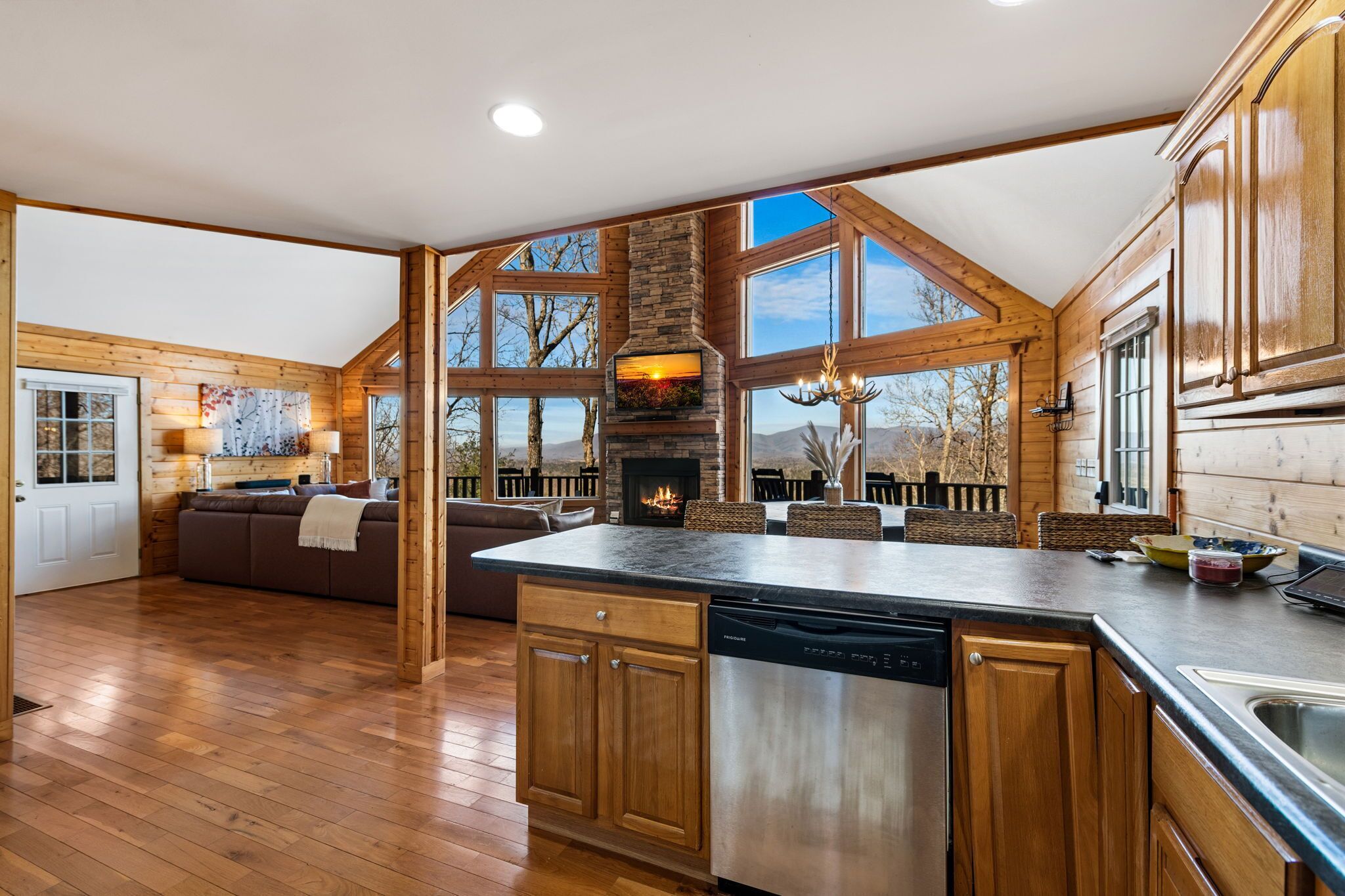 A spacious kitchen with a stone fireplace, wooden cabinets, and large windows offering views of the outdoors.