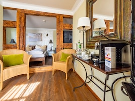 A hotel room with a wooden floor, a bed with green pillows, a wicker chair, a desk with a coffee maker, and a large mirror.