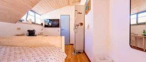 Ein Schlafzimmer mit Holzboden, einem Bett mit gemustertem Überwurf, einem Fernseher, einem kleinen Tisch mit Wasserkocher und einer Tür zu einem Badezimmer.