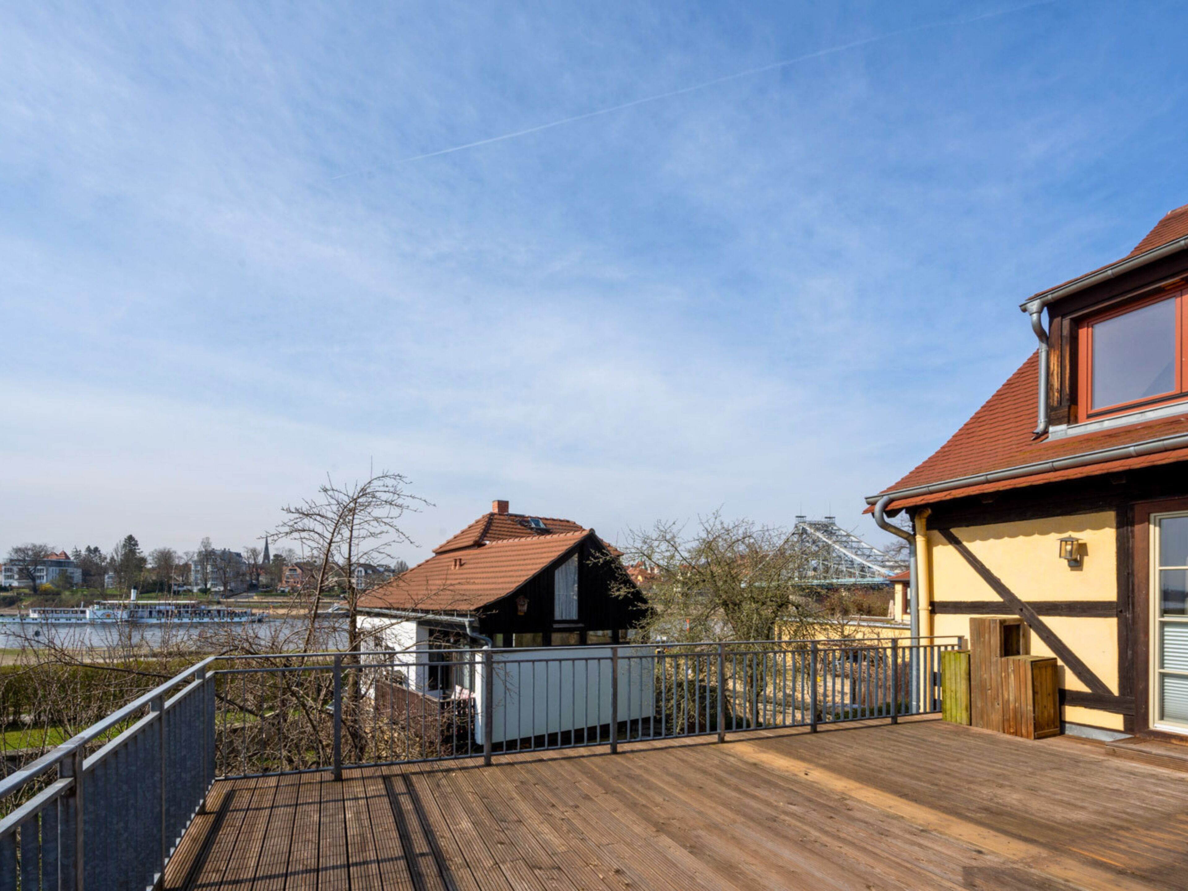 A wooden deck with a metal railing, a building with a red roof, and a view of a river and distant buildings.