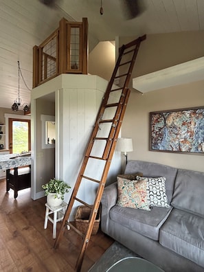 Ein Loft-Zimmer mit einer Holzleiter, einem grauen Sofa, einem gemusterten Kissen, einer Topfpflanze und einem Bild an der Wand.
