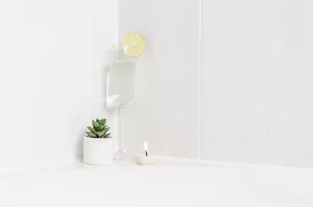 A minimalist room with a white wall, a small potted plant, a candle, and a glass of water with a lemon slice.