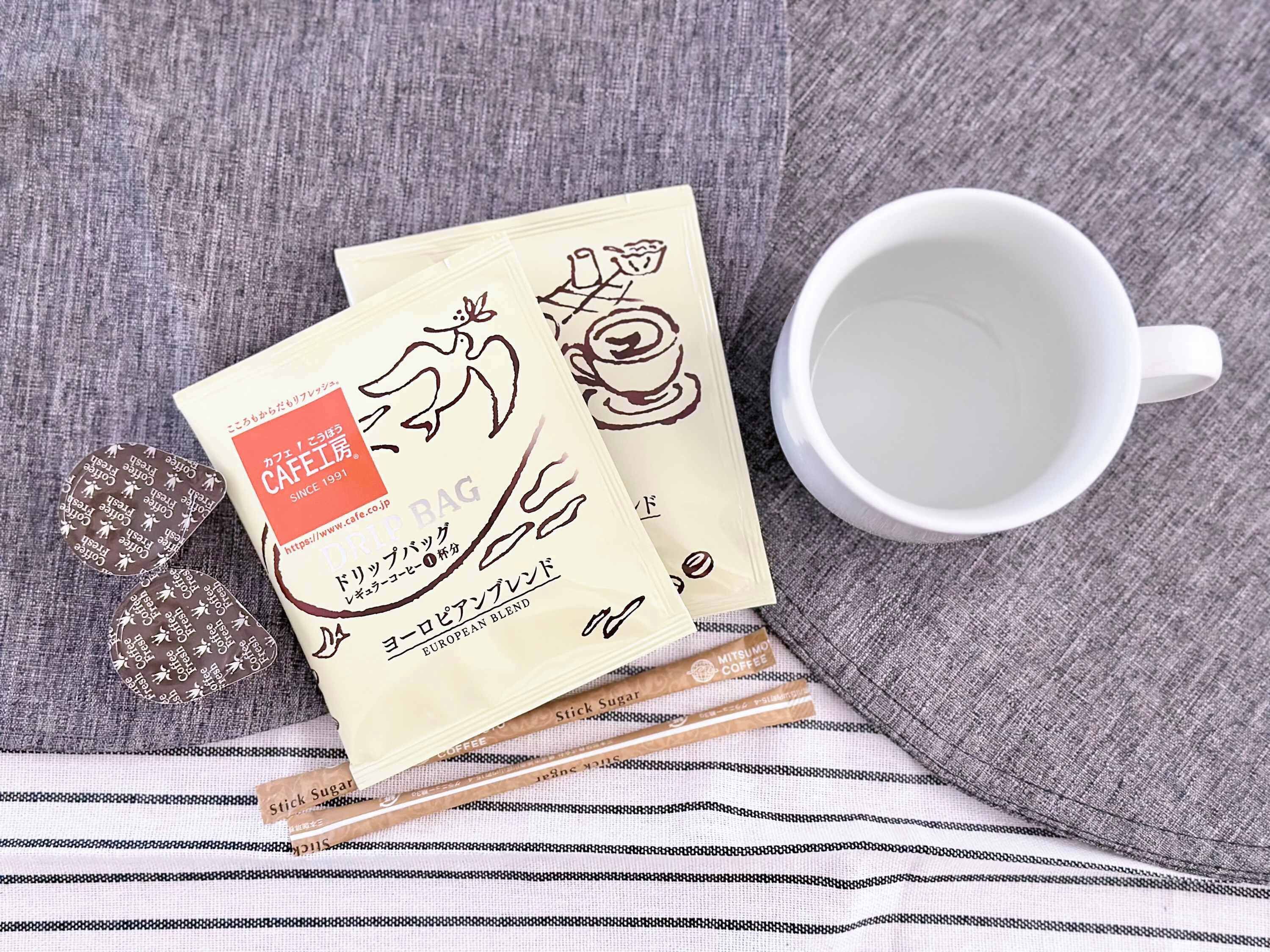 A white cup, two coffee bags, and a pair of chopsticks on a grey fabric surface.