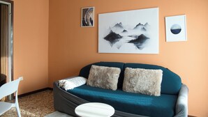 A room with an orange wall, a teal sofa with two fluffy pillows, a white chair, and two framed artworks depicting mountains and a moon. - Garden - Pino Mugo - Two Bedroom Apartment, Sleeps 6 (Barzio)