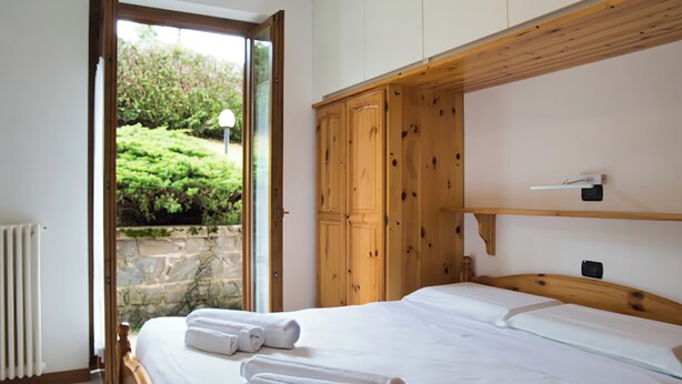 Un dormitorio con un armario de madera, cama con ropa de cama blanca y vistas a la vegetación a través de un ventanal.