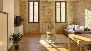 A spacious room with wooden floors, a sofa, a table, and two windows with wooden frames. - La Calade - Charming Apartment (Nîmes)