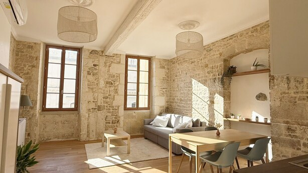 A rustic room with exposed stone walls, a wooden dining table with chairs, a sofa, and a small table with a lamp. - La Calade - Charming Apartment (Nîmes)