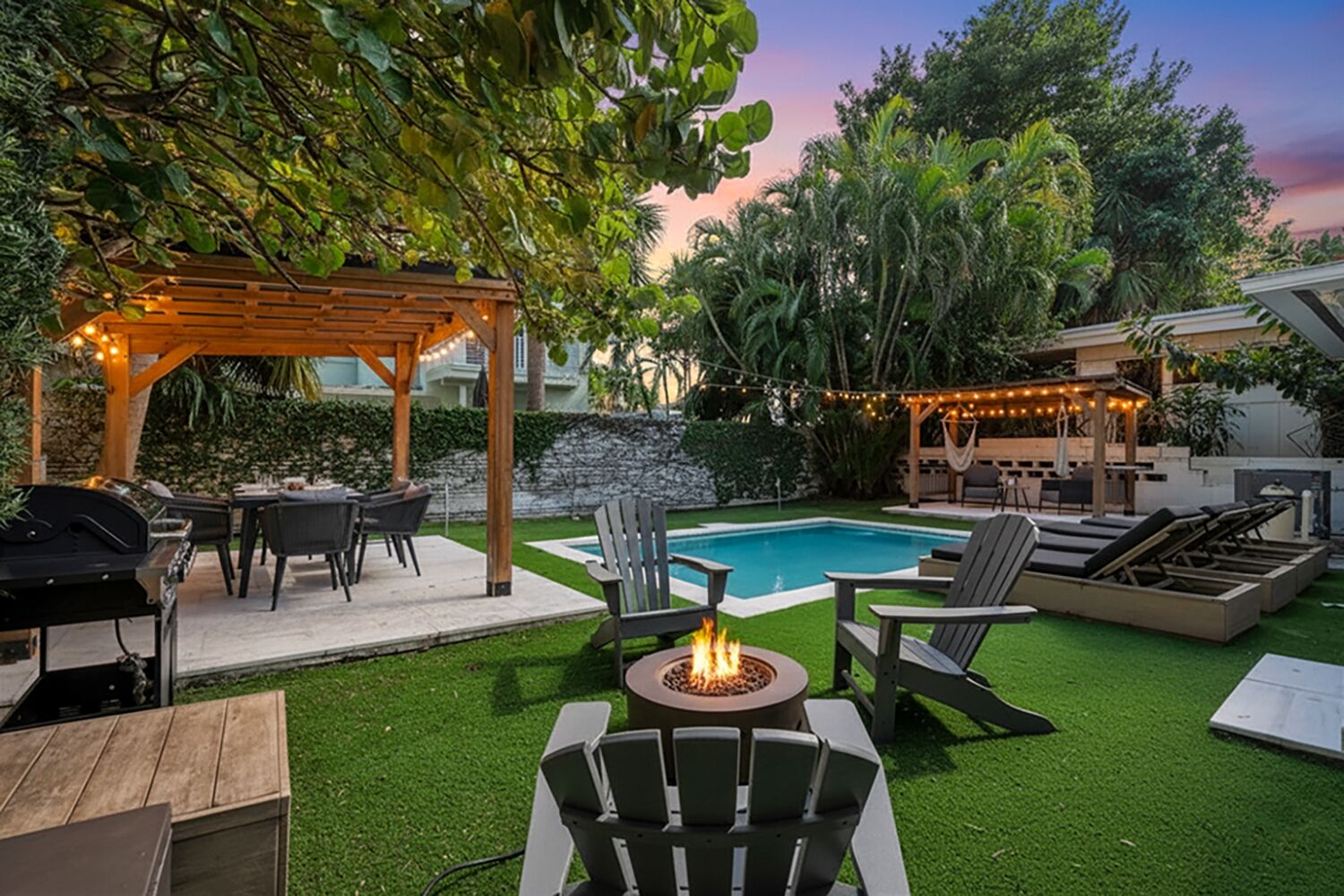 A backyard with a pool, outdoor seating, a pergola with string lights, and a fire pit.