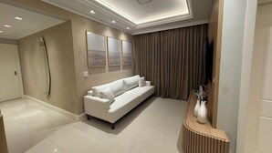 A modern living room with a white sofa, a wooden console table, and a large window with curtains. - Luxurious beachfront apartment in Itapema, SC. (Itapema)