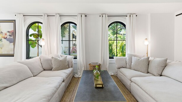 Ein modernes Wohnzimmer mit zwei weißen Sofas, einem zentralen Couchtisch und großen Fenstern mit weißen Vorhängen.