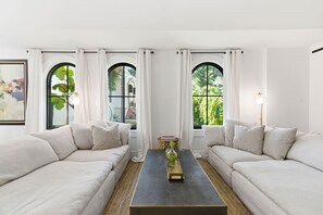 Una sala de estar moderna con dos sofás blancos, una mesa de centro y ventanas amplias con cortinas blancas.
