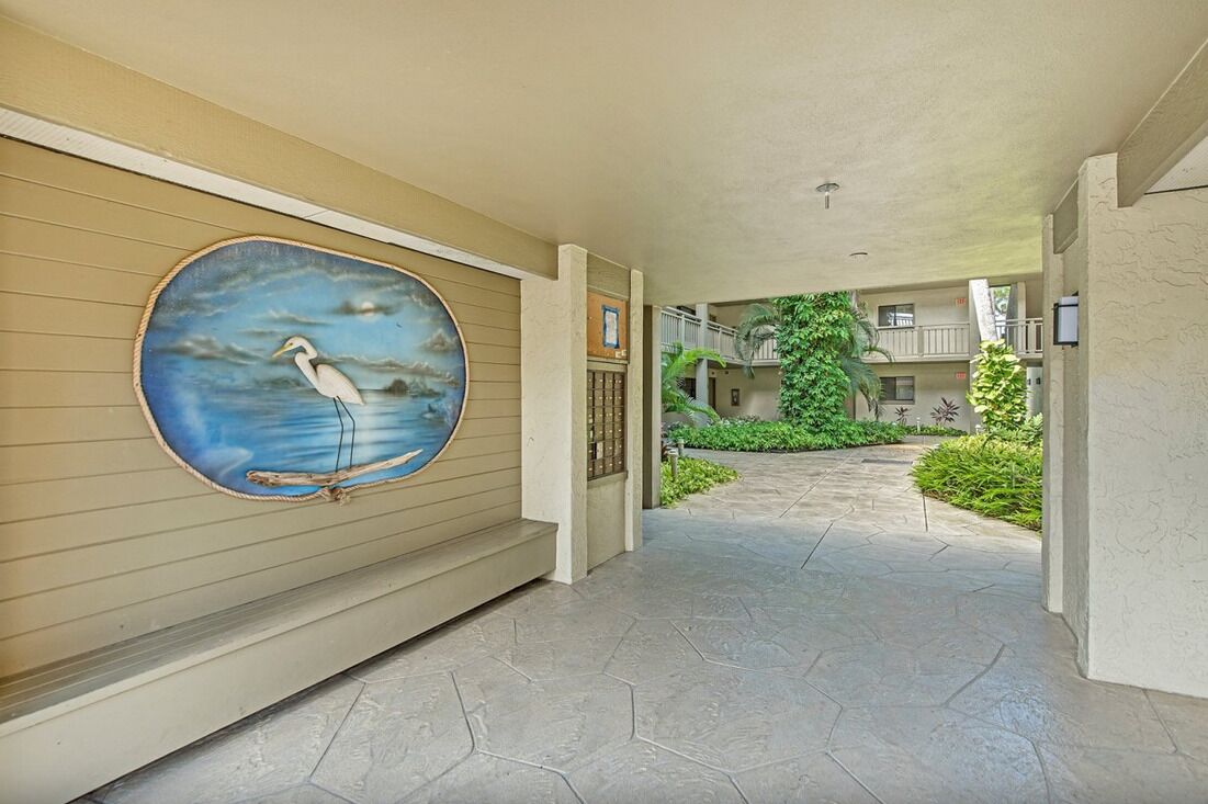 A corridor with a mural of a bird on the wall, a tiled floor, and a view of a building with balconies and greenery.