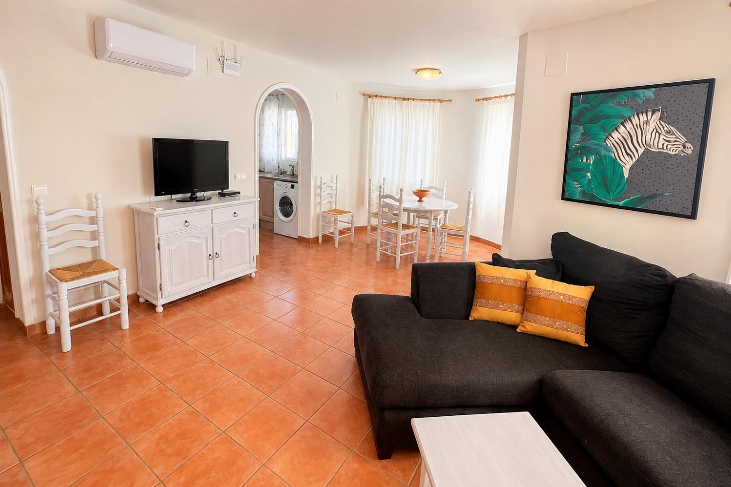 Een woonkamer met een zwarte bank, een witte kast, een televisie en een eethoek met een tafel en stoelen.