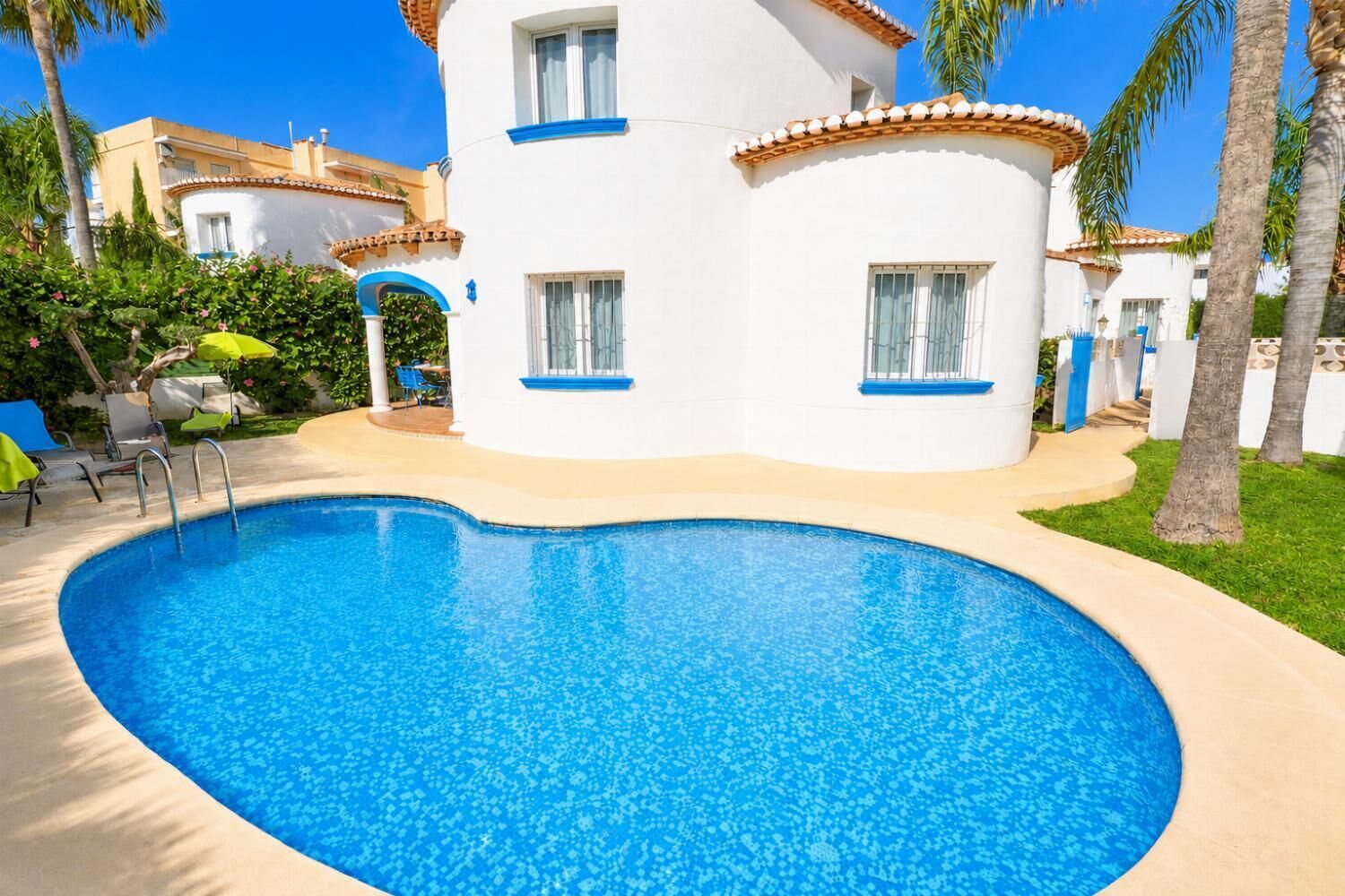 Una piscina con bordo curvo, circondata da un edificio bianco con decorazioni blu e finestre, e palme sullo sfondo.