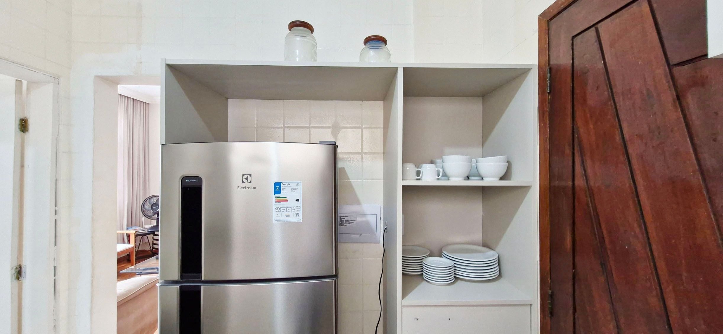 Een compacte keuken met een roestvrijstalen koelkast, een klein plankje met potten en een kast met gestapelde borden.