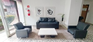 Ein modernes Wohnzimmer mit einer schwarzen Ledersofa, zwei Sesseln und einem weißen Couchtisch. An der Wand befinden sich zwei gerahmte Kunstwerke, ein Feuerlöscher und ein kleiner Topfpflanze.