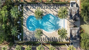 Eine Vogelperspektive zeigt einen Swimmingpool, der von Liegestühlen und Palmen umgeben ist, mit einem kleinen Gebäude auf der rechten Seite.
