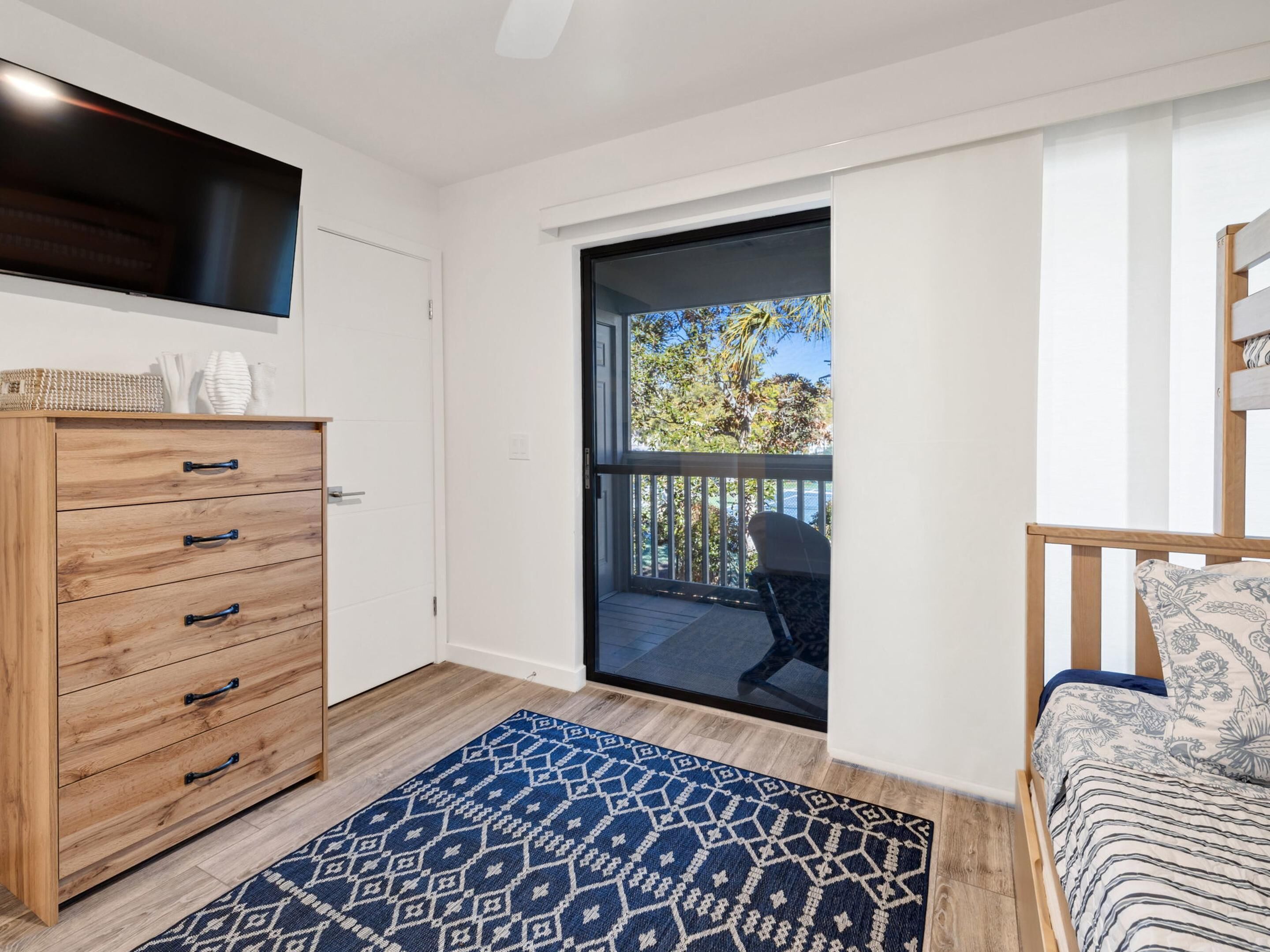 A bedroom with a wooden dresser, a patterned rug, a bed with a floral bedspread, and a balcony with a view of trees.