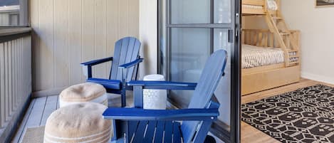 A balcony with two Adirondack chairs, a small round ottoman, and a wooden bunk bed with a patterned bedspread.