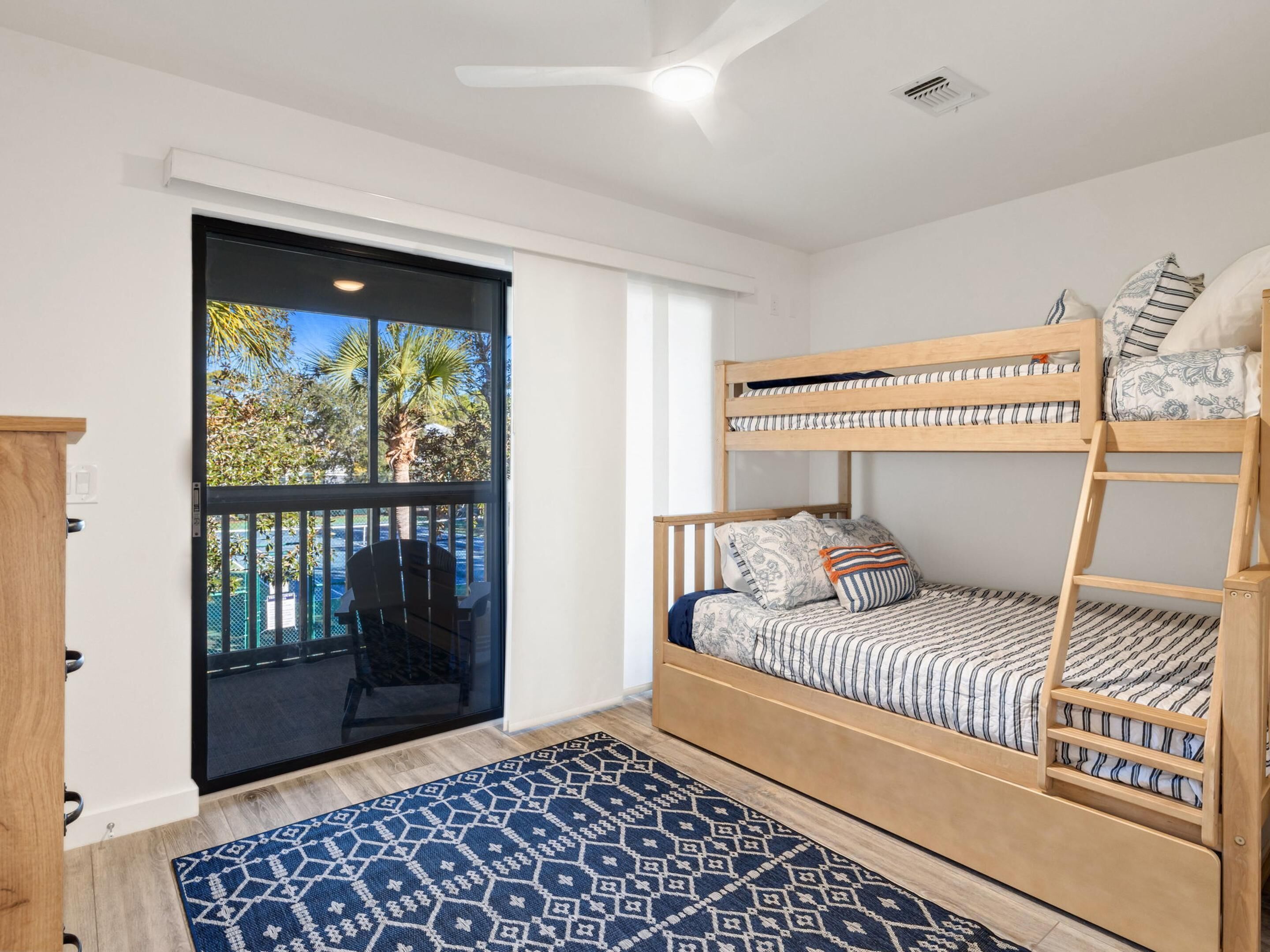 A room with a bunk bed, a chair, a patterned rug, and a view of palm trees through a sliding door.