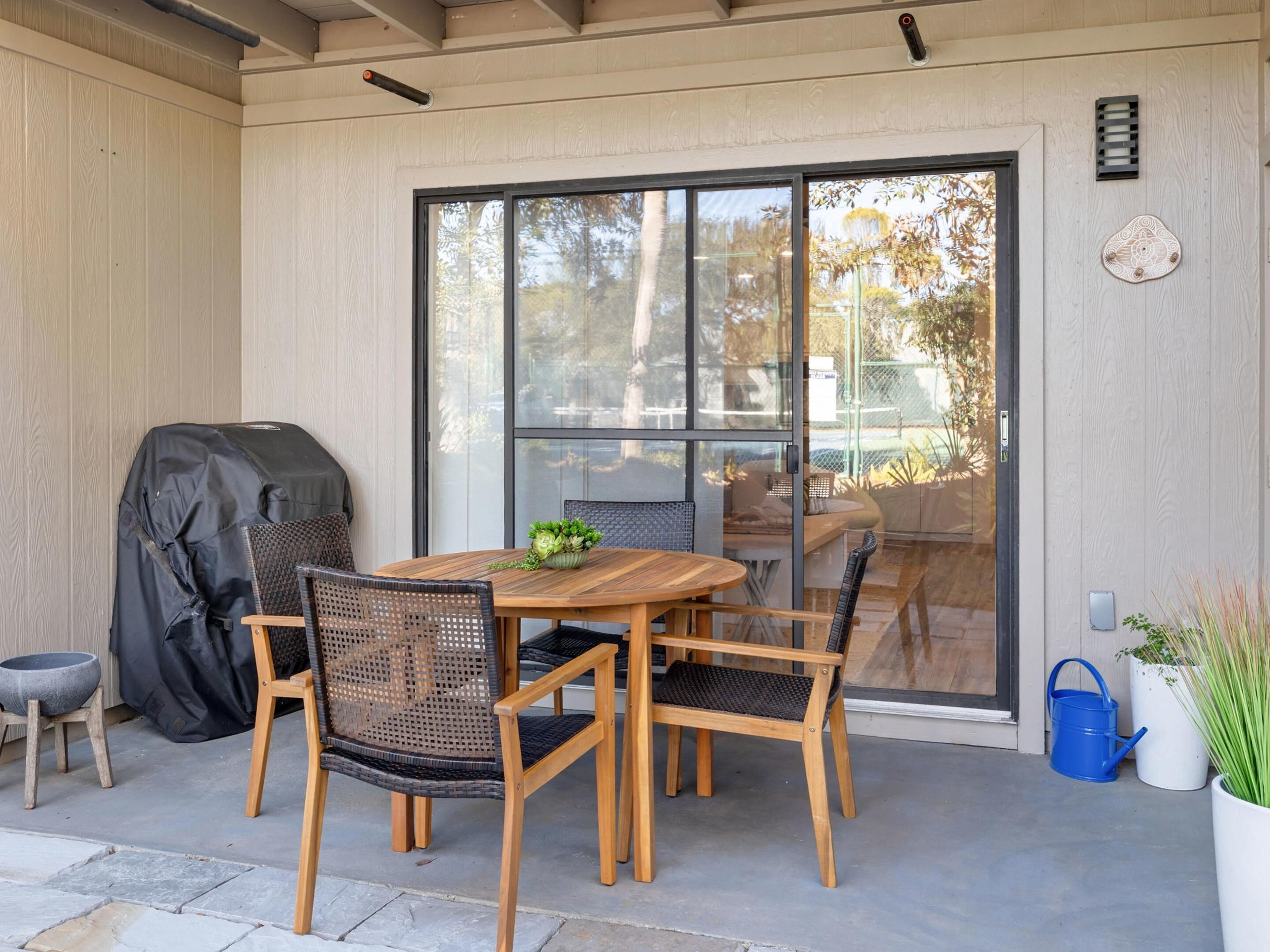Un patio exterior con mesa y sillas de madera, una parrilla y una puerta corrediza de vidrio que da a un jardín.