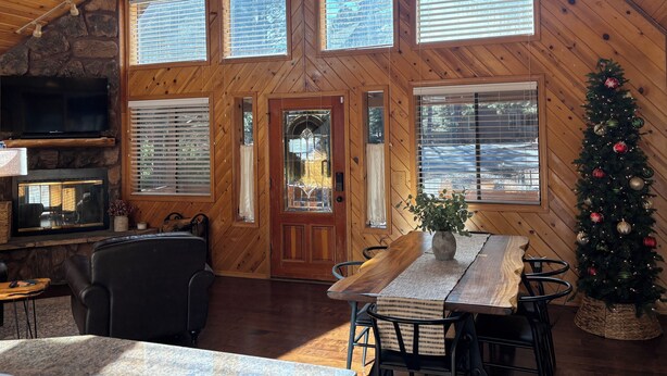 A cozy cabin interior with a fireplace, wooden walls, large windows, and a Christmas tree. - Christmas Tree Retreat – Cozy Forest Getaway in Pinetop - Sleeps 8 (Pinetop Country Club)