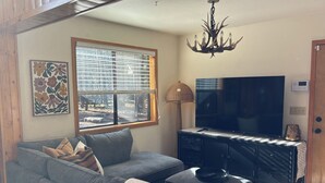 A living room with a grey sofa, a television, a chandelier, and a window with blinds. - Christmas Tree Retreat – Cozy Forest Getaway in Pinetop - Sleeps 8 (Pinetop Country Club)