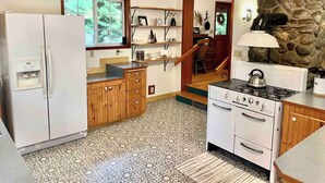 Una cocina con refrigerador, estufa, armarios de madera, pared de piedra y ventana con vista a la vegetación.