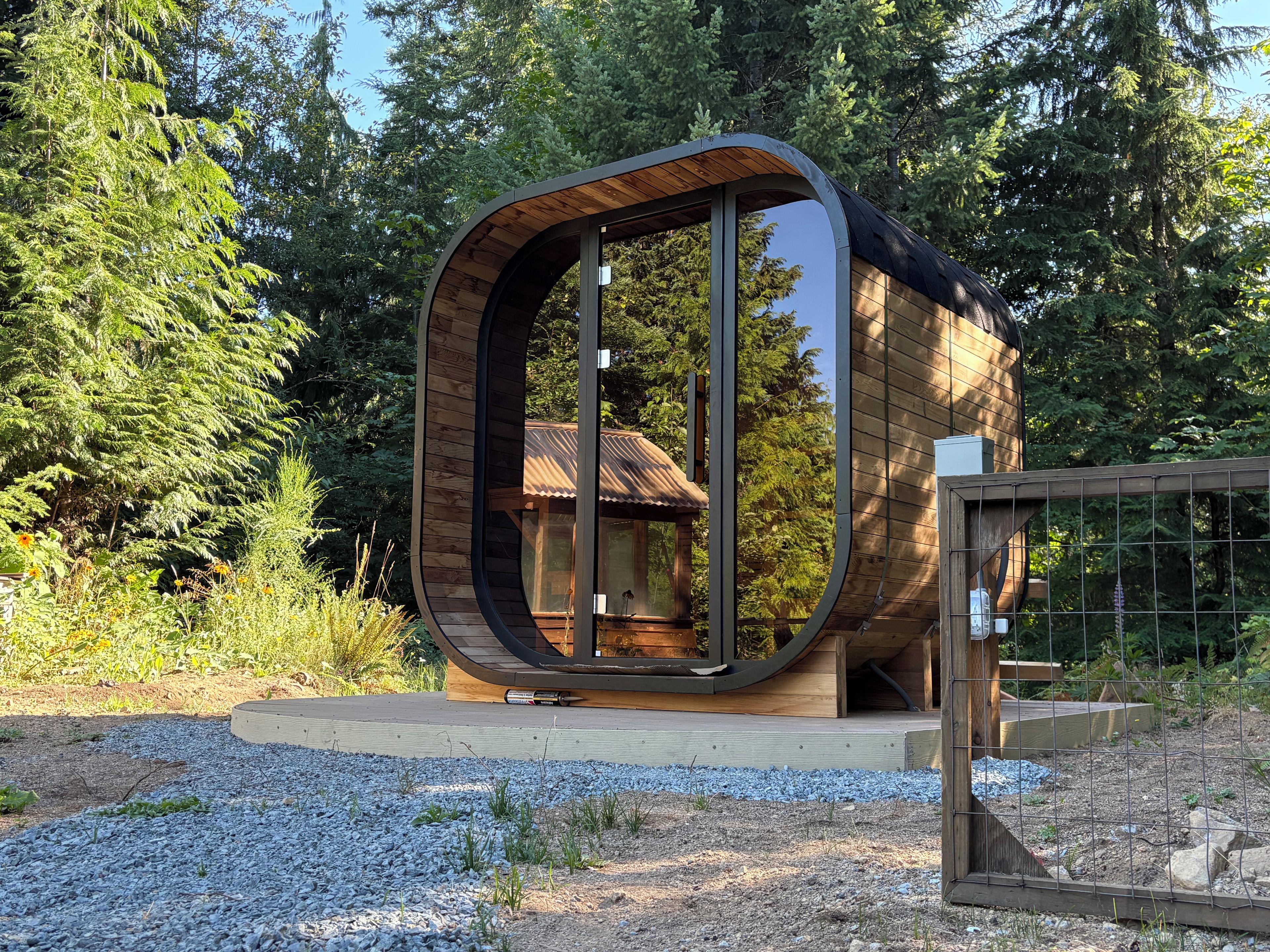 A modern, curved wooden cabin with a glass entrance, surrounded by trees and gravel.