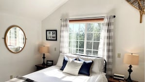 Un dormitorio con una cama grande, dos mesitas de noche, una mesita auxiliar, un espejo redondo y una ventana con cortinas.