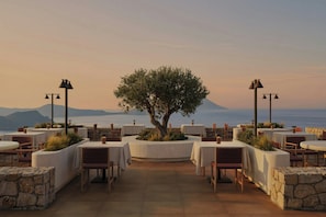 An outdoor dining area with round tables, chairs, and a central tree, overlooking a body of water and distant mountains. - Erema, a Member of Design Hotels (Milos)