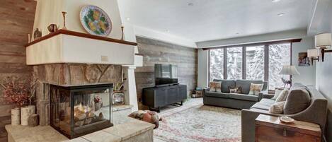 A modern living room with a stone fireplace, a television, a grey sofa, and a large window offering a view of snow-covered trees.
