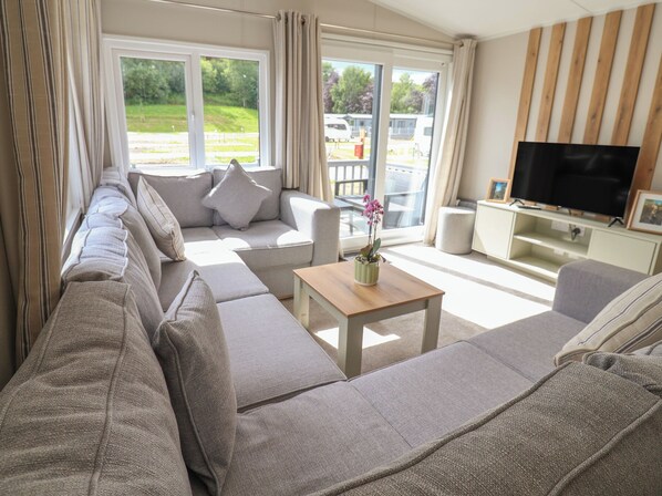 A modern living room with a sectional sofa, a TV, and a window with a view of a park. - The Oaks 2 Bedrooms (Northwich)