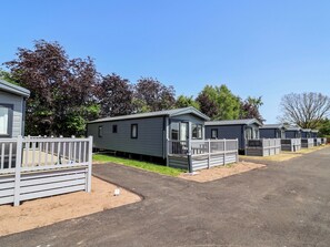A row of mobile homes with a fenced driveway, a clear sky, and trees in the background. - The Oaks 2 Bedrooms (Northwich)