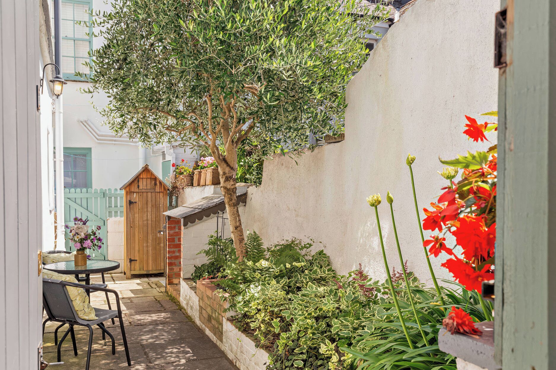 A narrow outdoor corridor with a wooden gate, a small table with chairs, and a variety of plants and flowers.