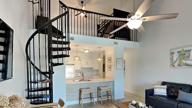 A spacious living area with a high ceiling, a black spiral staircase, a ceiling fan, a kitchen area, and a seating area with a sofa and chairs. - Treasure Cove #d308 - Paradise City (Panama City Beach)