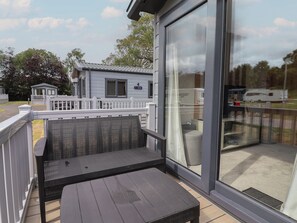 A covered outdoor seating area with a table and chairs, a white railing, and a view of a mobile home in the background. - The Oaks 3 Bedrooms (Northwich)