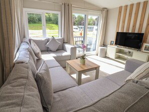 A modern living room with a sectional sofa, a TV, and a window with a view of a park. - The Oaks 3 Bedrooms (Northwich)