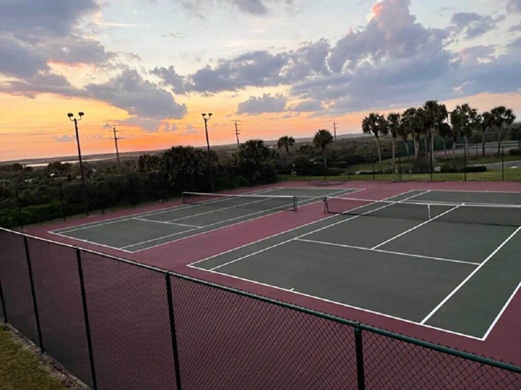 An outdoor tennis court with a red and green surface, surrounded by a chain-link fence and palm trees.